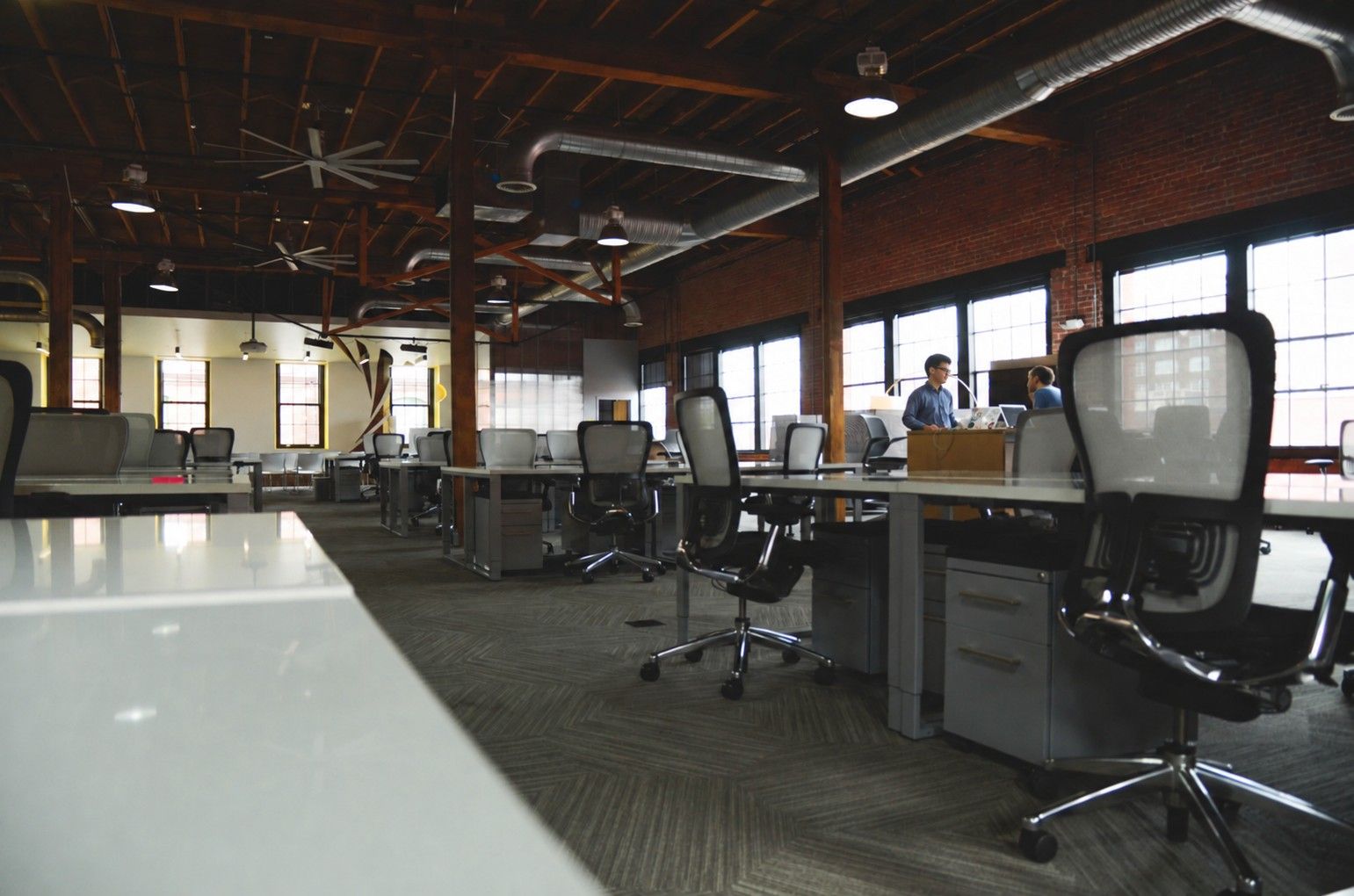 An open-plan office with rows of desks, two people talking by the windows